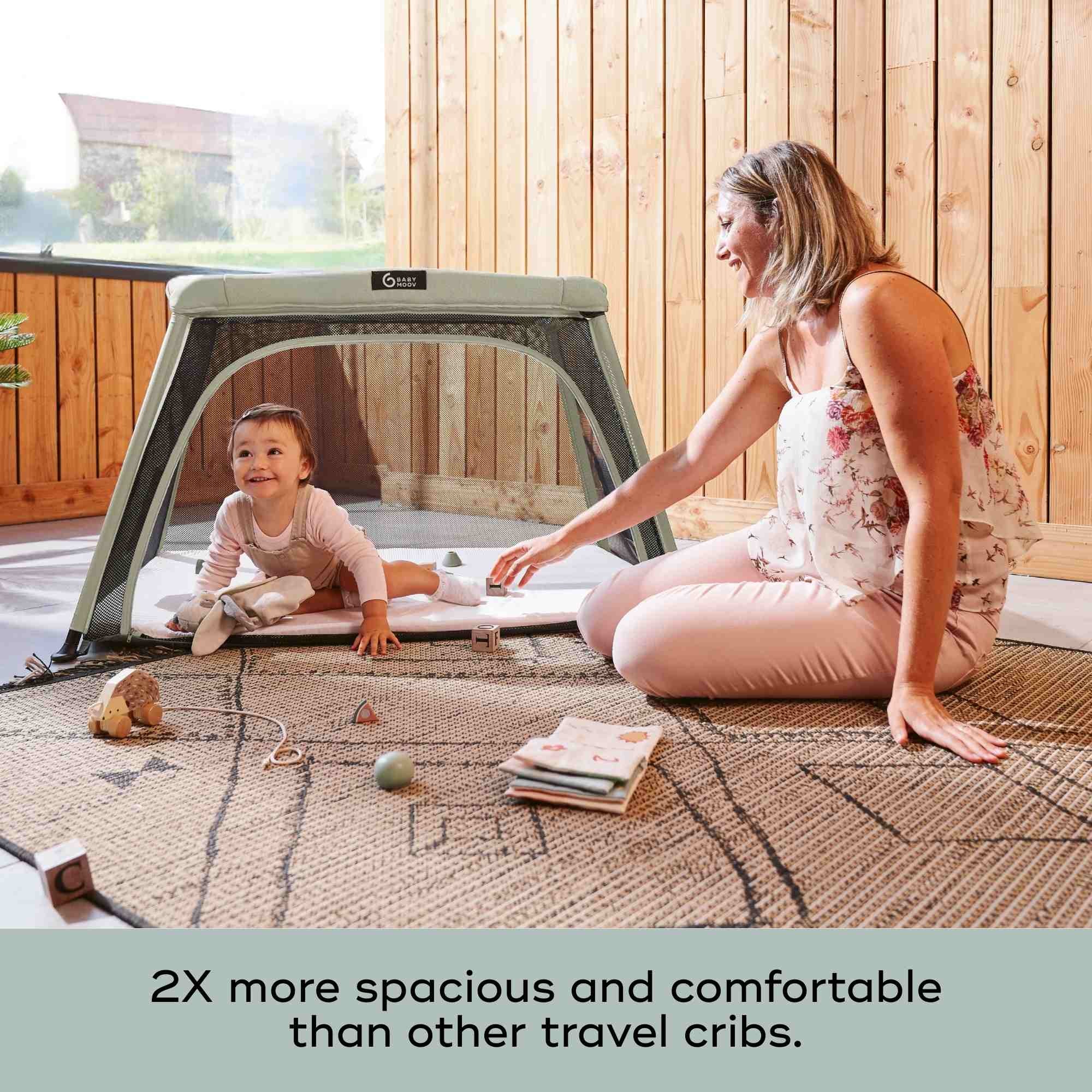 Woman and child playing on a spacious travel crib in a wooden room.